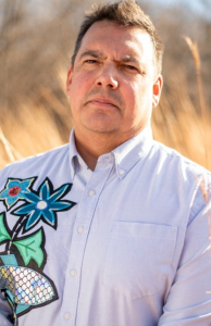 A man poses in front of tall grass. He is wearing a button-up shirt with large flowers on the front
