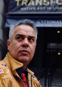 A man wearing la leather coat and adorned with beaded plants and a beaded necktie looks up and to his right