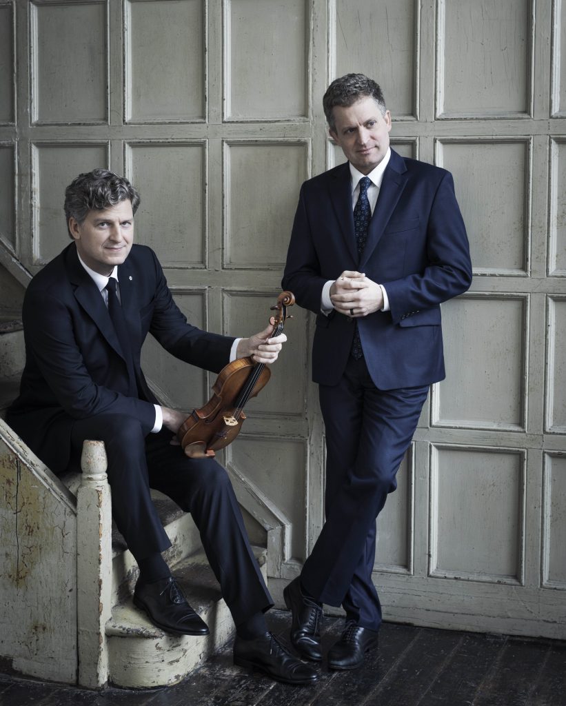 Two men in suits: One sits on stairs, holding a violin in his knee, while the other man stands