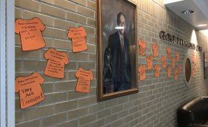 A portrait hangs from a brick wall, surrounded by orange shirts cut out of construction paper, with messages written on them.