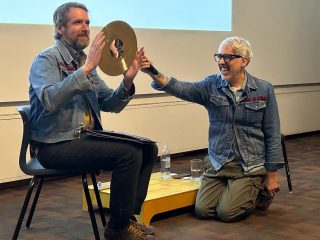 A man sits an a chair and holds a cymbal, while another man reaches toward him with a microphone