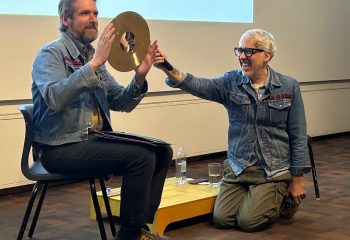 A man sits an a chair and holds a cymbal, while another man reaches toward him with a microphone