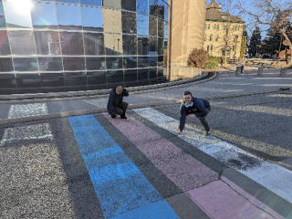 Two people crouch over a painted rainbow crosswalk, using chalk to cover tire marks on it.